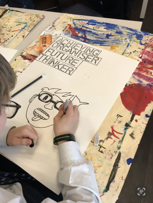 A student sketches a cartoon on a paper at a cluttered art table strewn with colorful paint marks. Beside the drawing, a paper with 'ACHIEVING ORGANISER FUTURE THINKER' suggests a creative educational setting.
